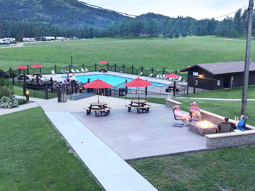 Four people relax around a fire pit on a patio with a pool in the background.