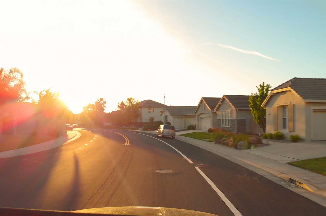 POV LENS FLARE: Driving through the scenic streets of a rich neighborhood on the fringes of San Francisco at sunset. Evening sunbeams shine down on people's luxury homes. Idyllic suburbs at sunrise.