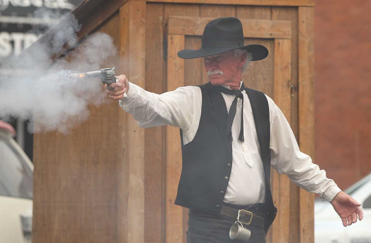 A man with silver hair in Western regalia fires a pistol in a cloud of smoke