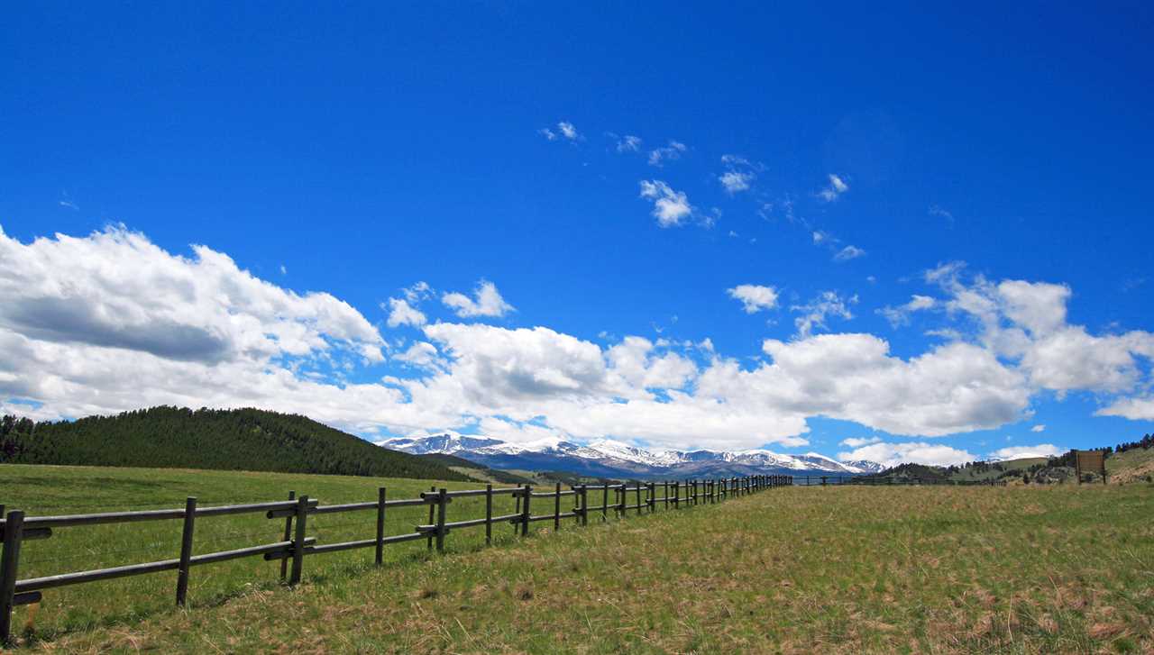 A wooden fence cuts across a sprawling field fringed by hills under a blue sky.