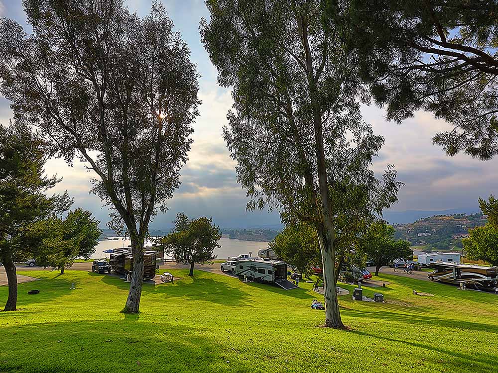 RVs parked near a grassy slop under tall trees with a lovely lake in the background.