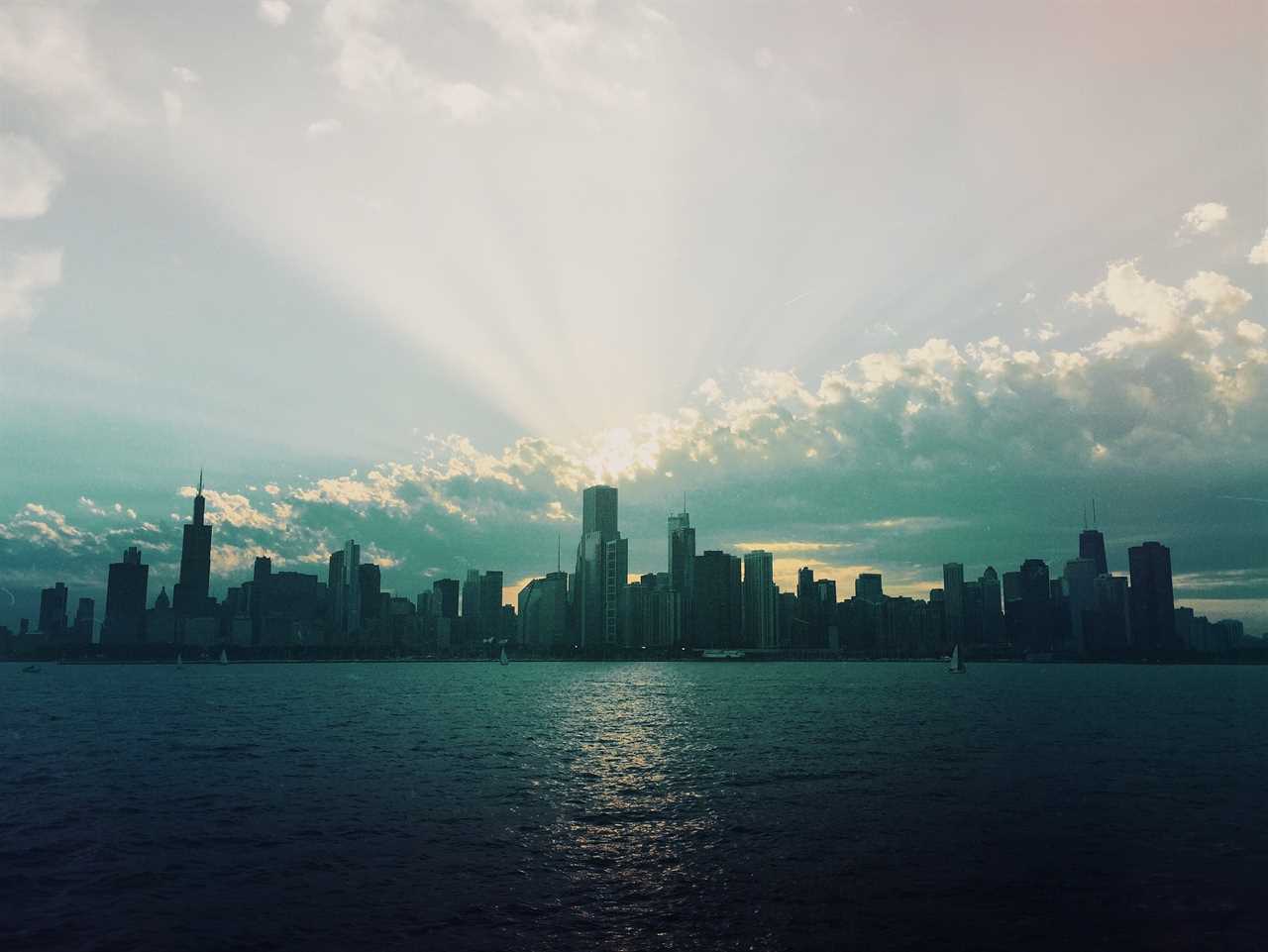 Sun sets behind a thicket of towering sky scrapers that loom over water.