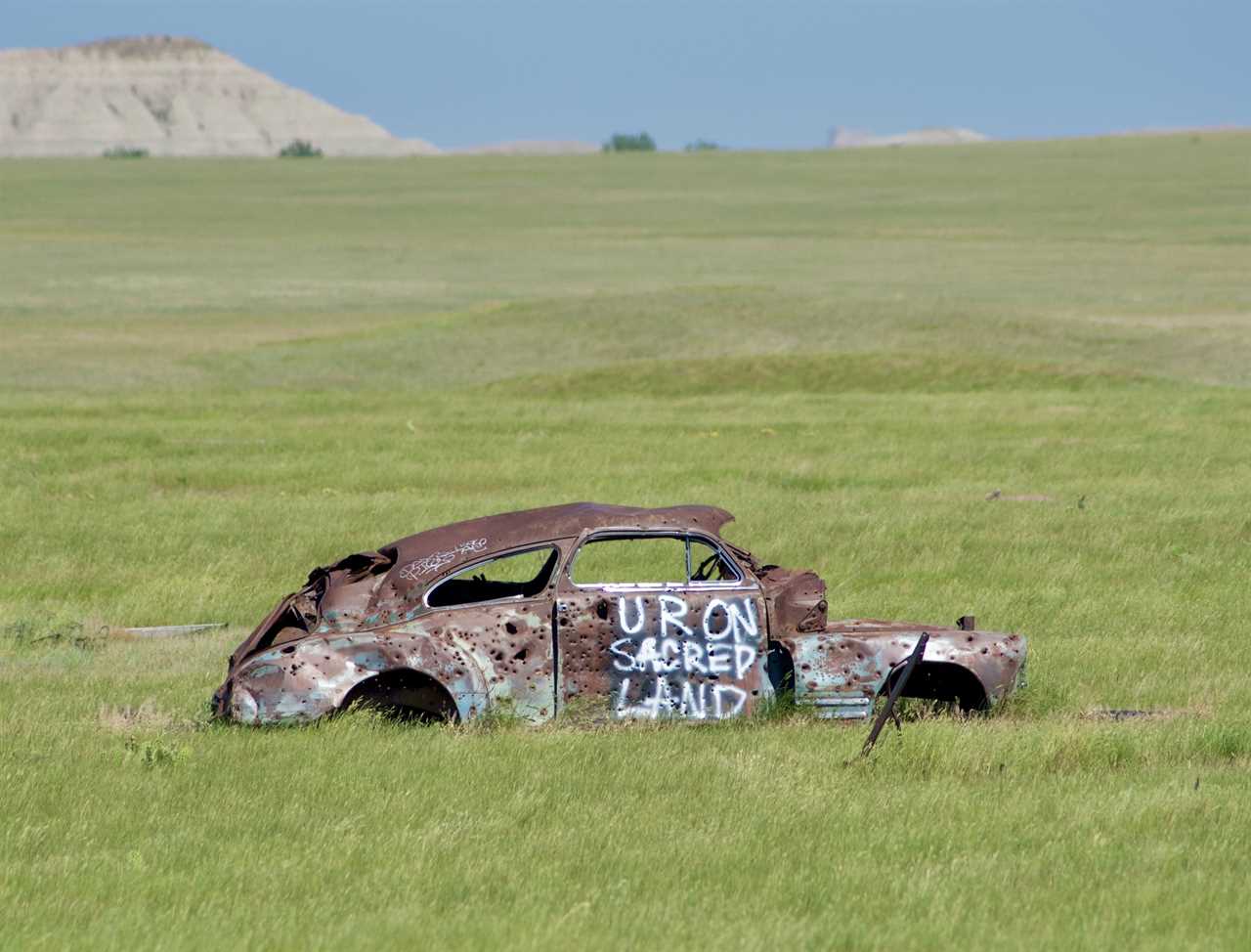 A rusted VW bug with "U R On Sacred Land" painted on it.