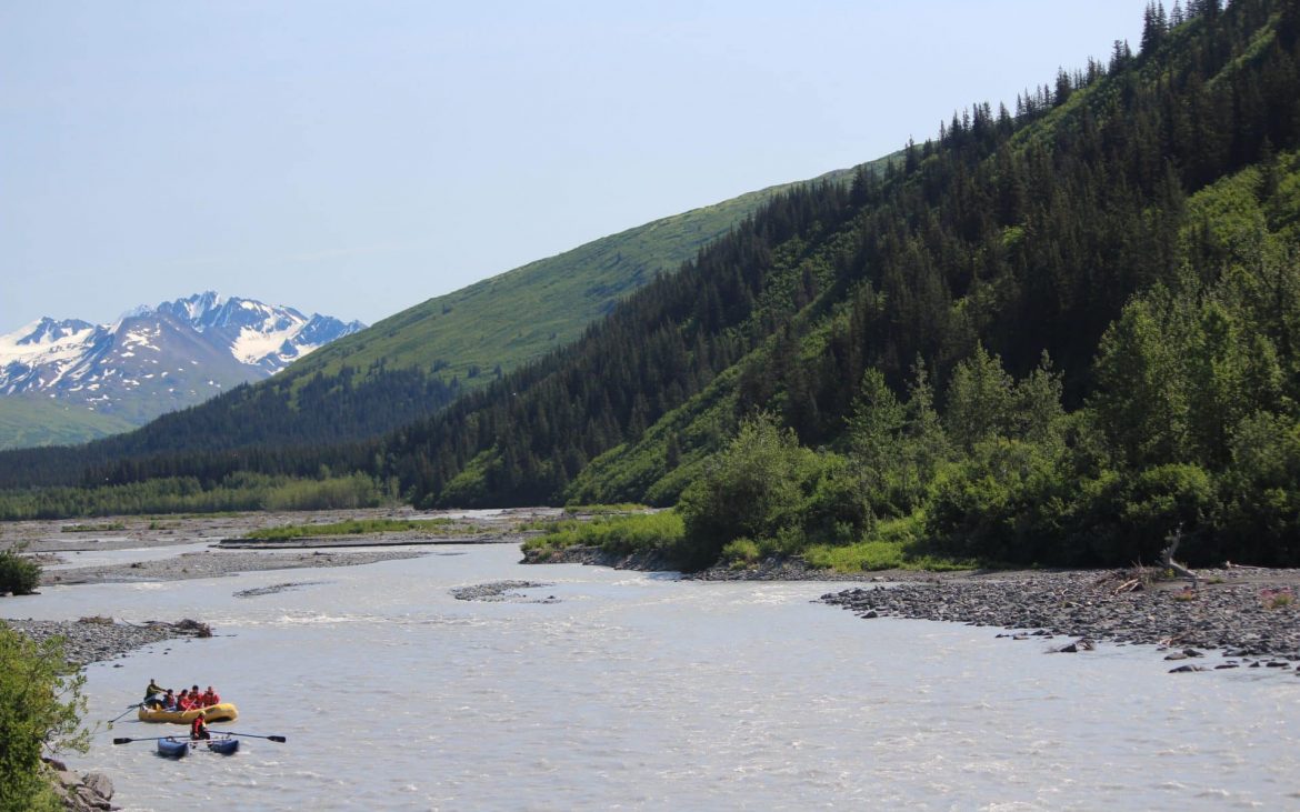 Raft full of people white water rafting down Alaskan river with trees in background