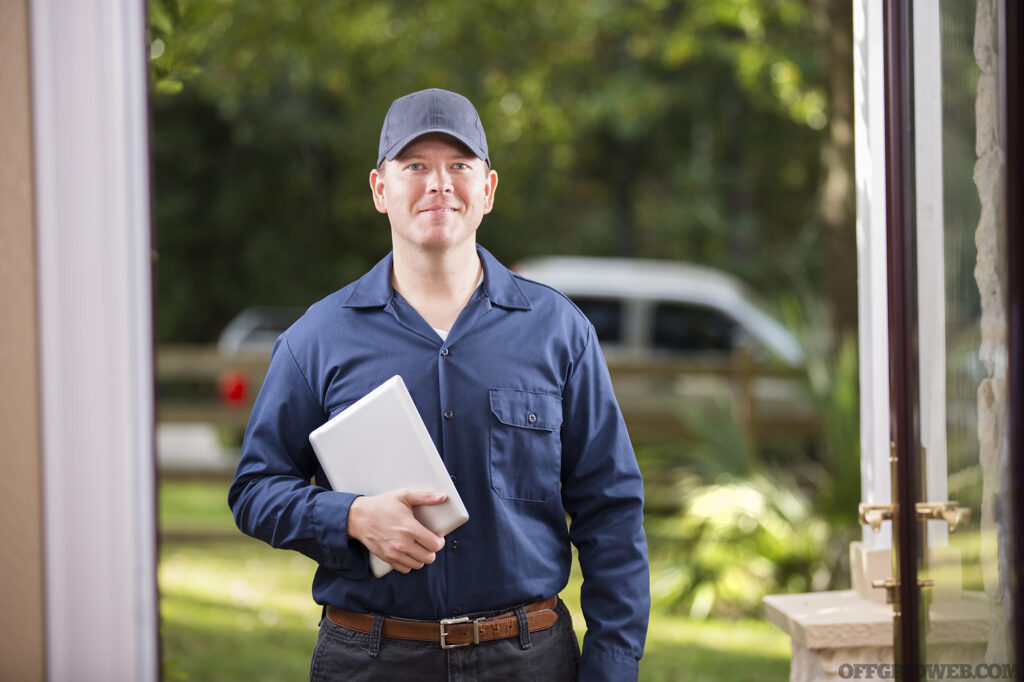 Photo of a middle aged adult repairman or blue collar/service industry worker makes service/house call at customer's front door. He holds his digital tablet with work invoices. Inspector, exterminator, electrician. He wears a navy colored uniform. Service truck seen in background parked on road.