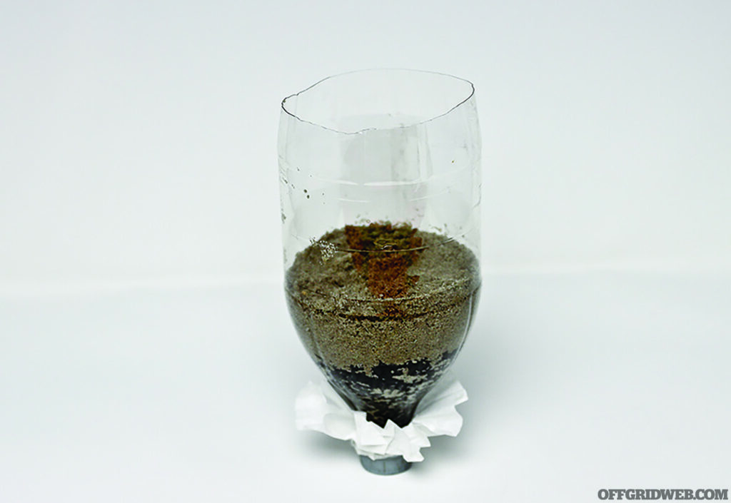 Photo of an upside down, clear plastic soda bottle with a layer of activated charcoal on the bottom, and a layer of undyed sand on the top.