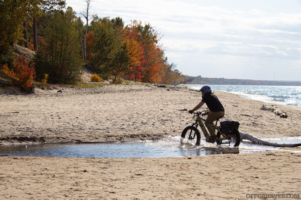 Photo of an adult male crossing a shallow river outlet on a fat-tire eBike.