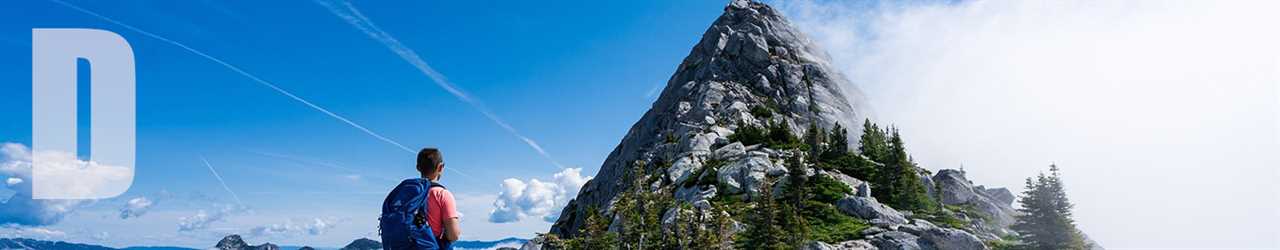 A backpacker hikes toward a steep mountain partly shrouded in cloud, careful to practice good camping etiquette.