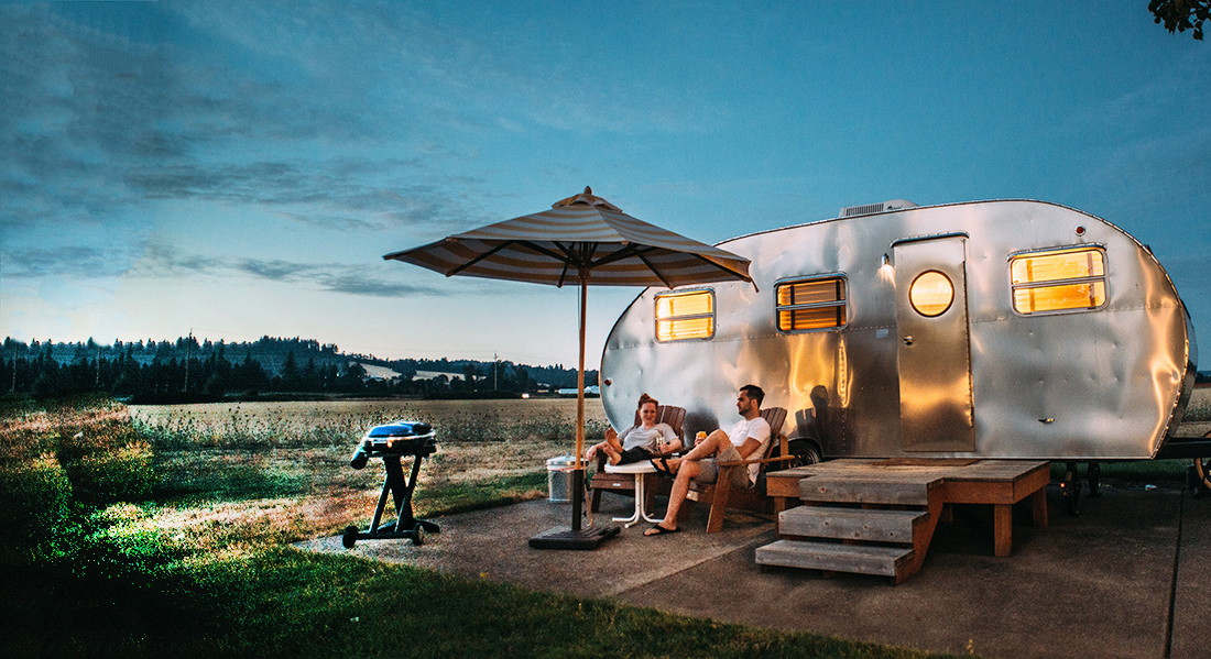 A couple sitting outside of a silver motorhome with grill.