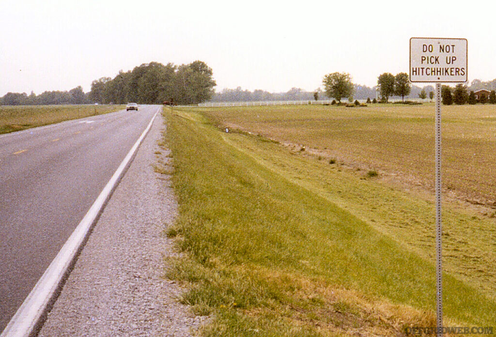 Photo of a sign on the side of a rural highway warning drivers against picking up hitchhikers.