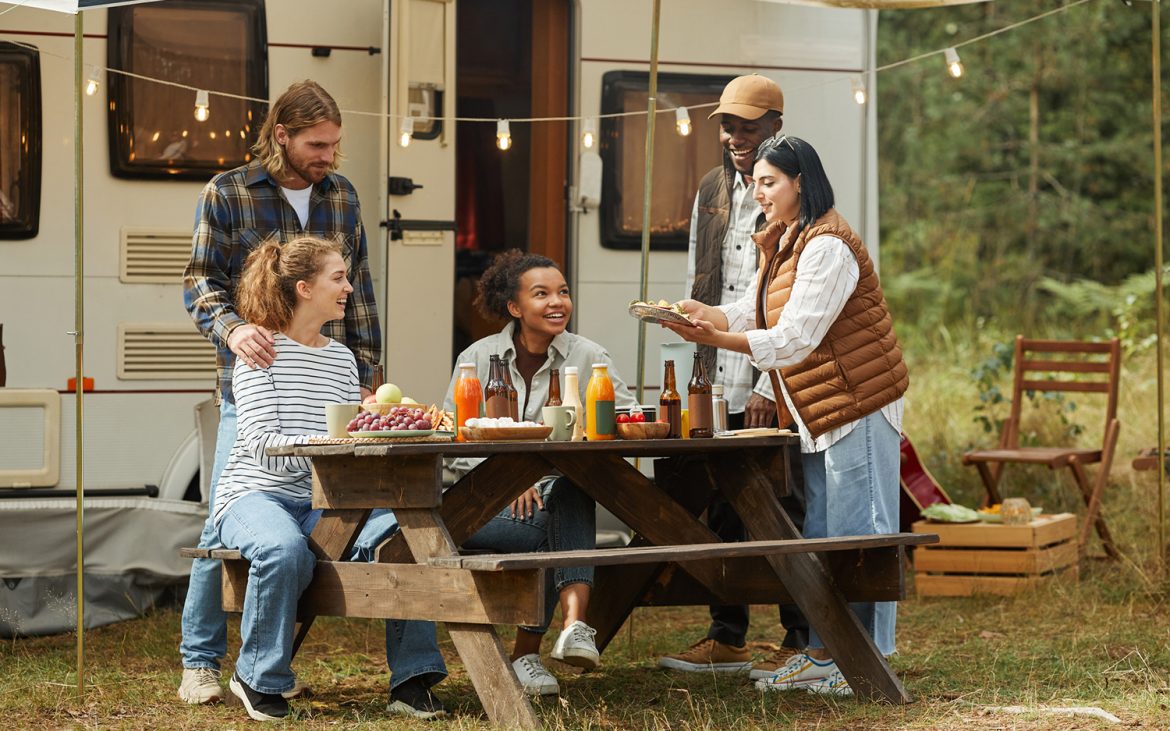 Full length view at diverse group of young people enjoying picnic outdoors while camping with trailer van