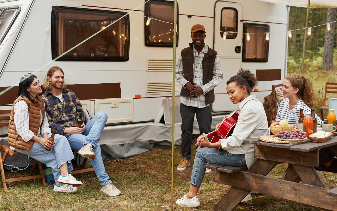 Full length portrait of young African-American woman playing guitar while enjoying camping outdoors with diverse group of friends