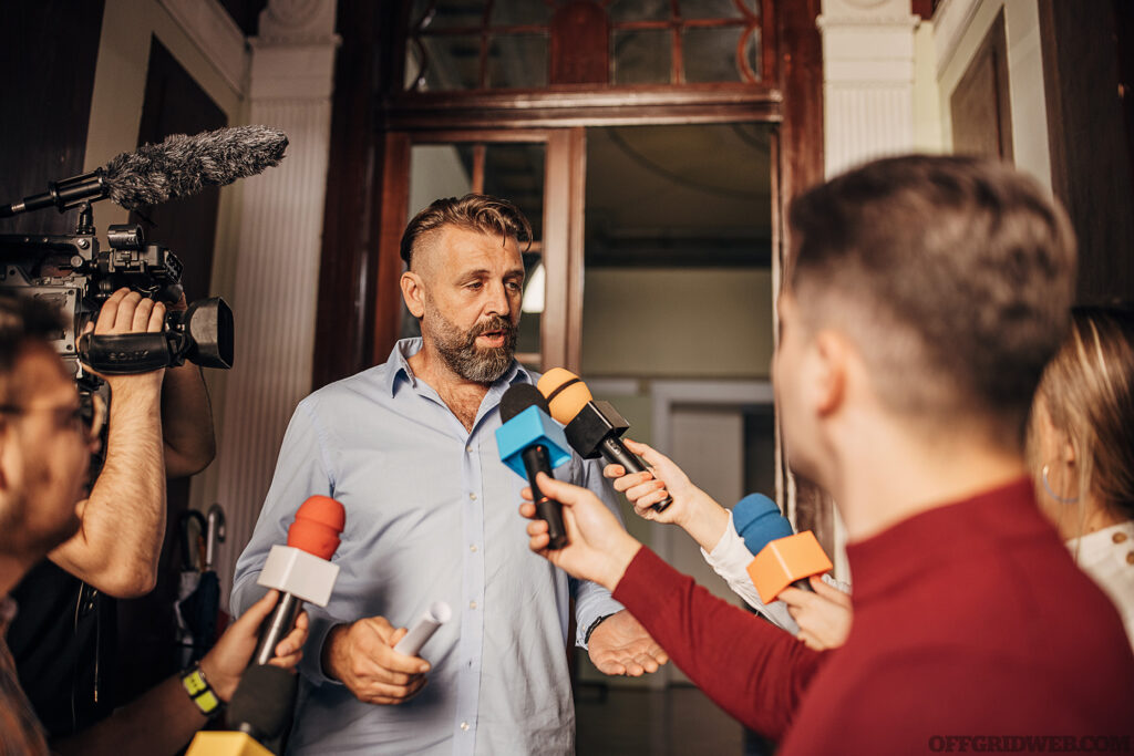 Group of people, male politician confronted by journalists with microphones.