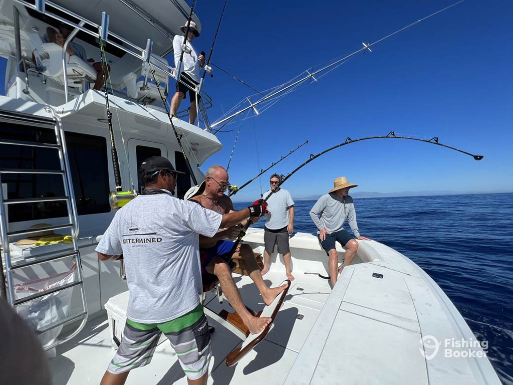 A view of a man helping another man sat in a fighting chair on the back of an offshore sportfishing boat in San Jose Del Cabo as he tries to battle it out with whatever is on the end of his heavy-duty line with two other people standing near the edge of the boat behind him