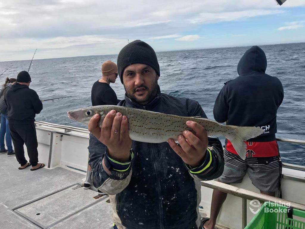 A man in a beanie hat holding a Whiting fish towards the camera aboard a fishing charter in Perth on a cloudy day,. with a number of other anglers dressed for winter visible behind him