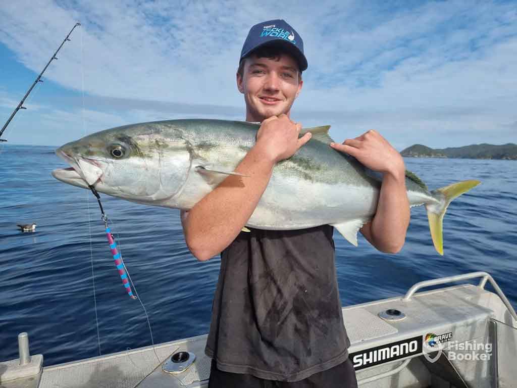 A late-teenage boy wearing a baseball cap with sunglasses on them holds a Kingfish with both arms aboard a fishing charter in Auckland on a sunny day with the water visible behind him