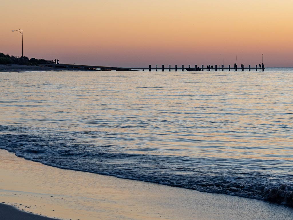 A view across a beach looking towards North Point Jetty in Perth at sunset, with the ocean water calmly brushing against the sandy beach