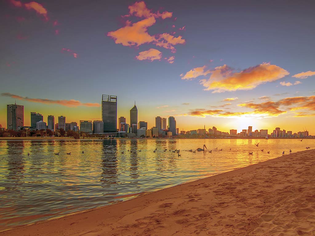 A view from a sandy beach across the Swan River towards Perth's skyline at sunset on a clear day