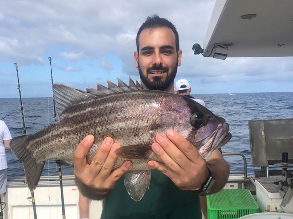 A man holding a Black Bream aboard a fishing charter in Perth on a summer's day, with the open waters of the ocean across the deck of the boat behind him