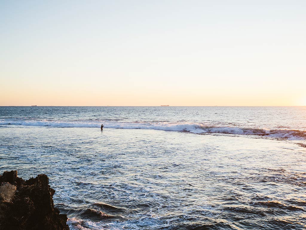 A view across the water from a rocky shoreline towards a lone angler wading and surf fishing on a clear day near sunset