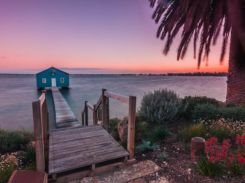 A view towards a boat house and small wooden dock on the Swan River near Perth at sunset, with a palm tree visible on the right of the image