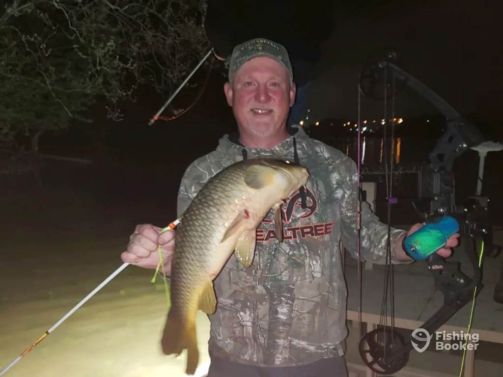 A bowfishing angler at night standing aboard a boat with the boat's lights visible, as he holds a Carp with the bow that caught it