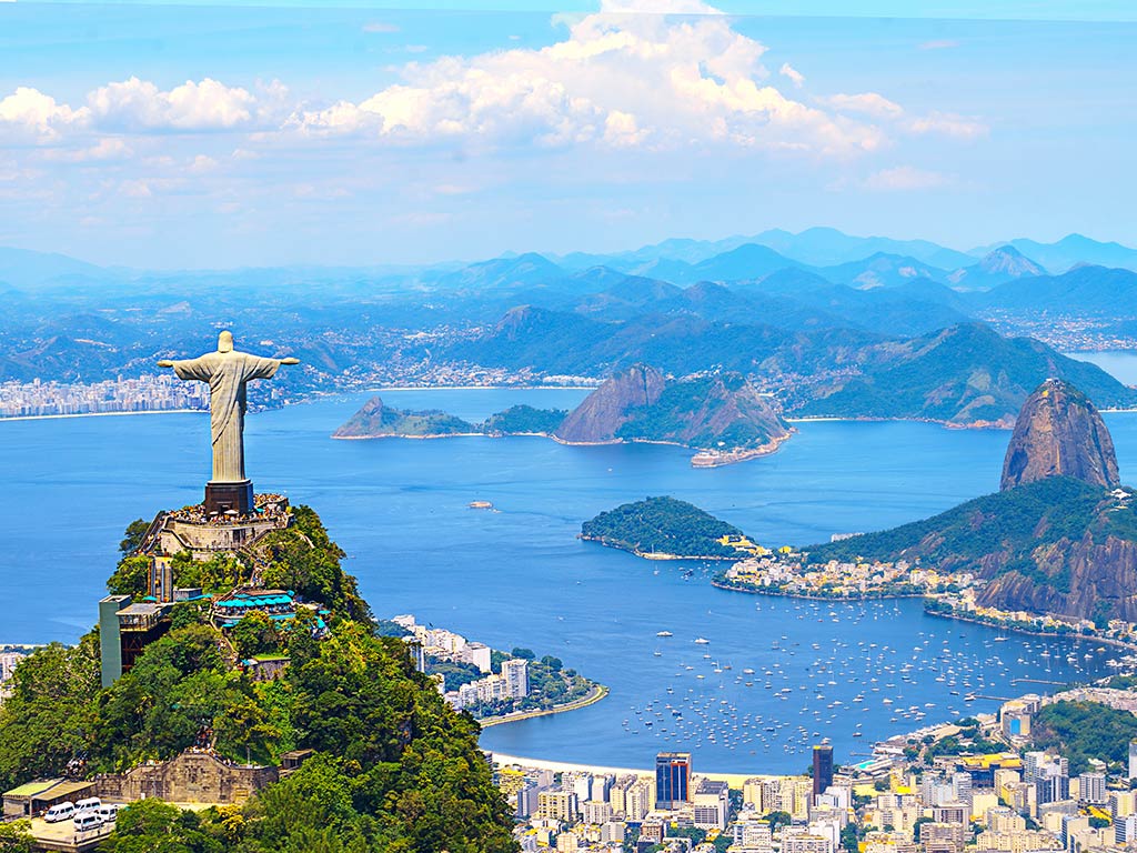 An aerial view of RIo de Janeiro on a bright day, with the Christ the Redeemer statue visible on the left of the image and the Atlantic Ocean visible in the distance