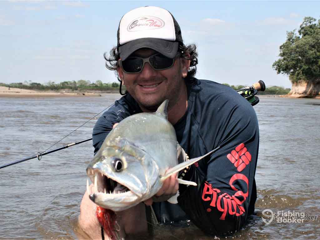 An angler in a baseball cap and sunglasses, wasding up to his waist in a river in Colombia on a clear day, and presenting his Payara catch to the camera