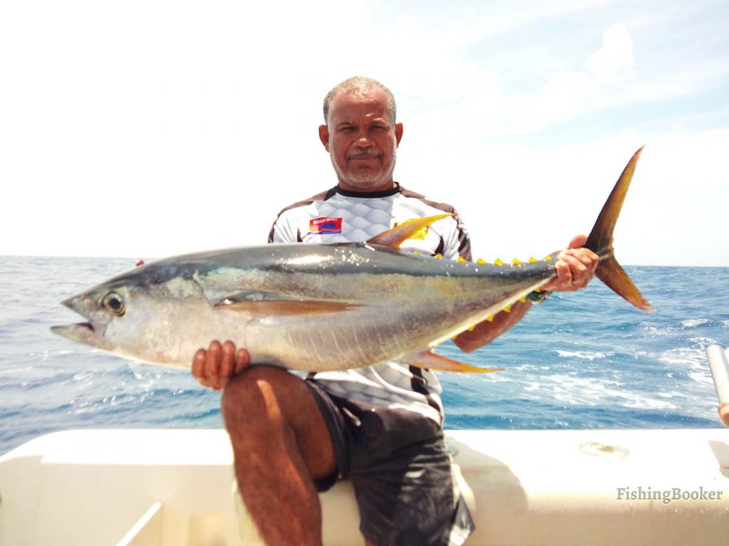 A man looking professional in fishing gear aboard the deck of a fishing charter on a clear day holding a large Tuna