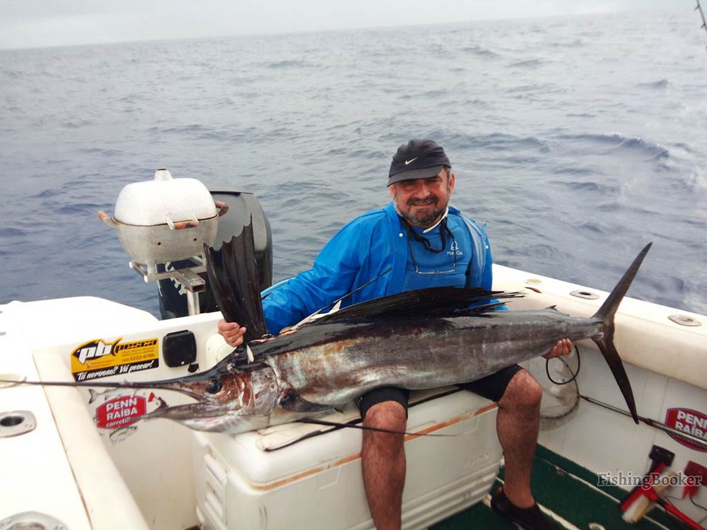 A man in a blue sweatshirt and baseball cap aboard a deep sea fishing charter in Brazil with a Sailfish laid across his lap on a cloudy day with the water visible behind him