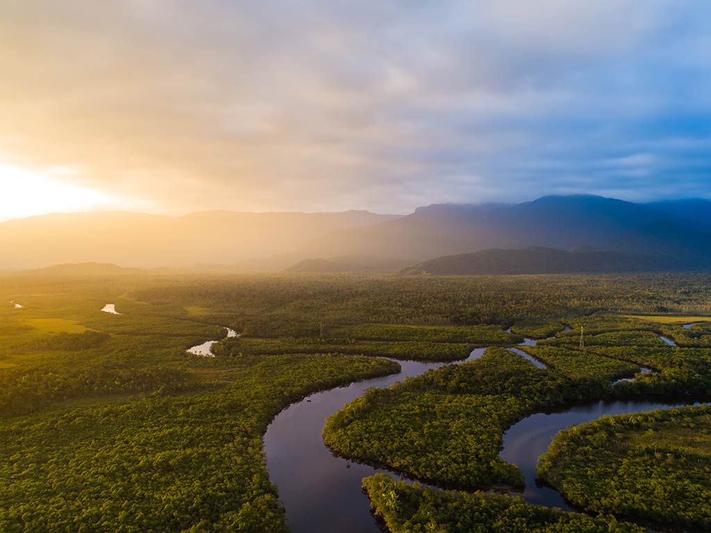 An aerial view of the Amazon river, winding its way throuigh the rainforest at sunset on a bright day, with some mountains visible in the distance