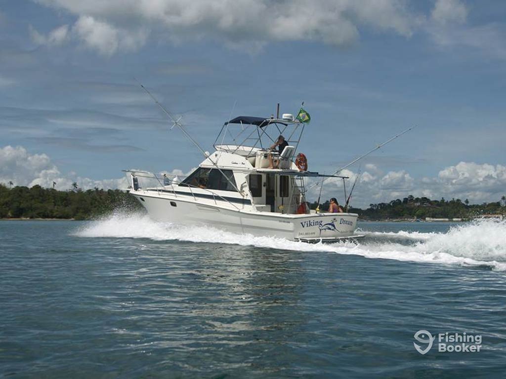 A view across the water towards a charter fishing boat in saltwater in Brazil on a sunny day, with a tree-lined shore visible in the distance