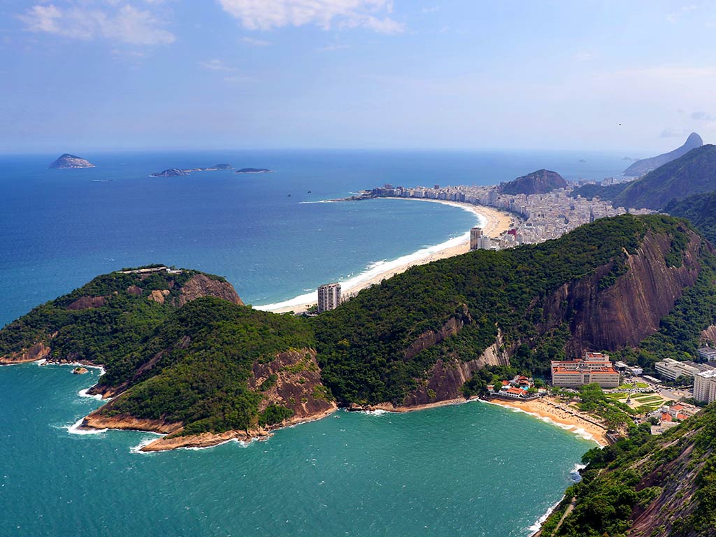 An aerial view looking towards the Copa Cabana Beach in Brazil on a clear day, with another smaller beach visible in the foreground before a peninsula sticks out, separating it from the Copa Cabana