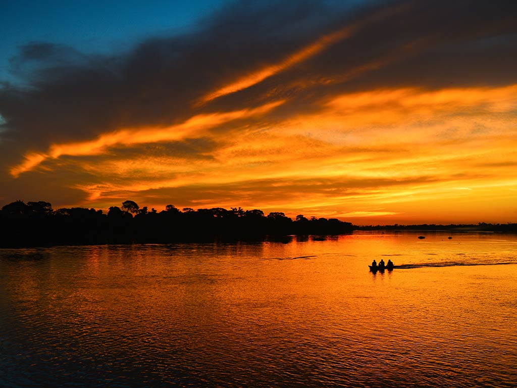 A view across the water of the Amazon in Brazil towards a fishing boat at sunset, with the orange sky in the distance and a silhouette of a boat visible on the water