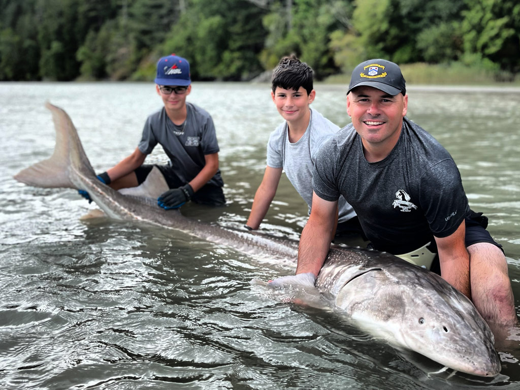 A photo of a father and his two sons squatting in a river surrounded by a forest while posing proudly with a Sturgeon so big that it could easily be a state fish record