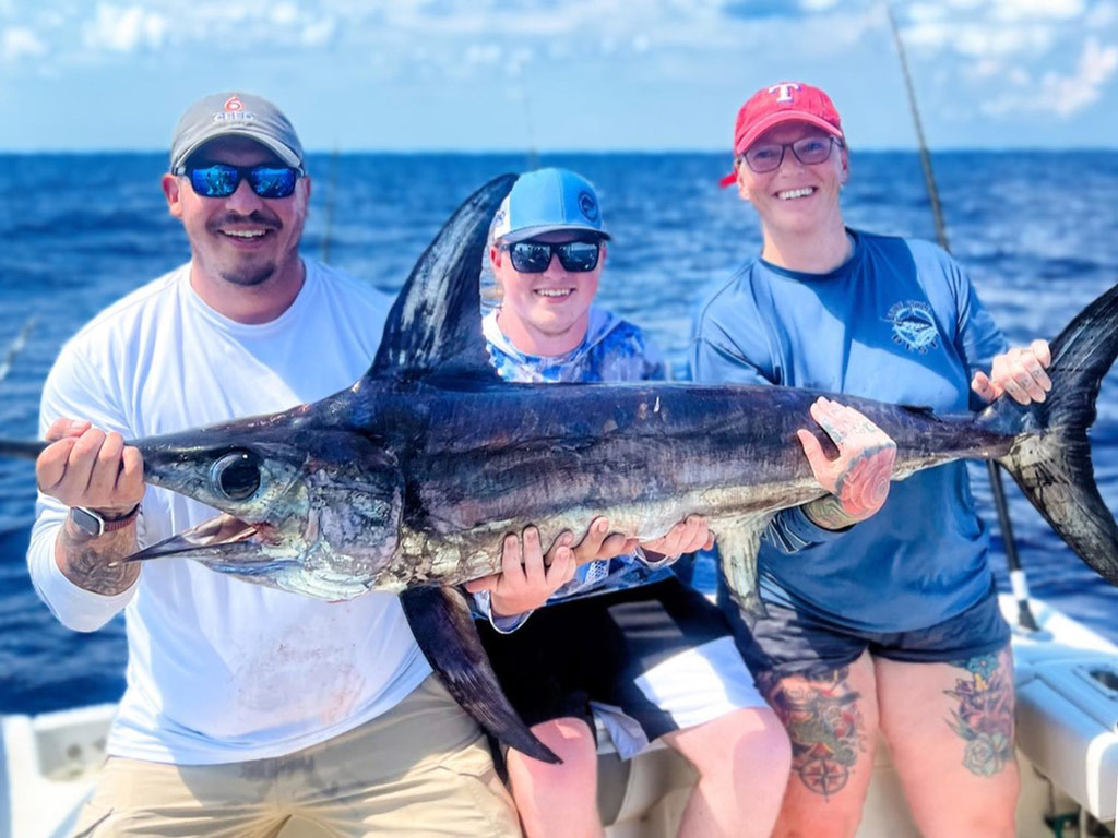 A group of three anglers sitting on a charter fishing boat and posing with a big Swordfish caught while fishing offshore on a bright and sunny day