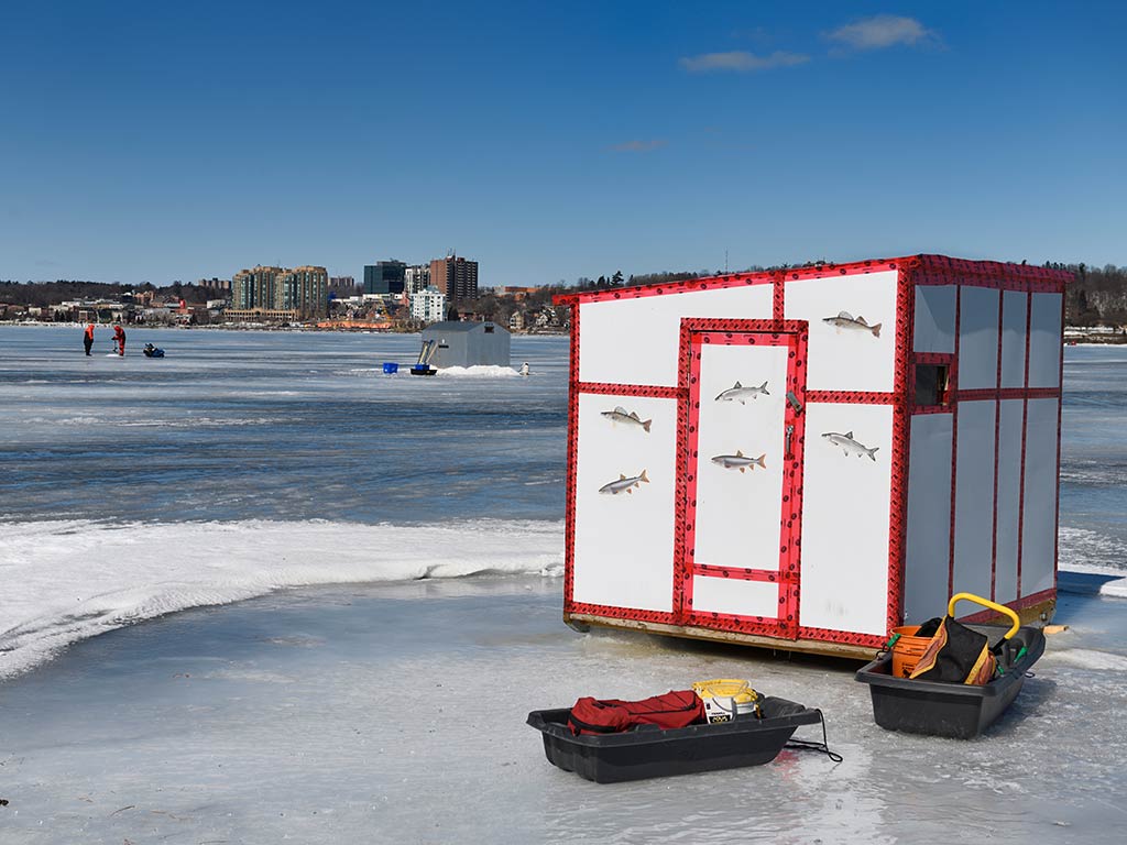 A view across the ice of Lake Simcoe on a clear winter's day towards a city in the distance with an ice hut and a couple of sleighs set up in the foreground with numerous others dotted around in the distance