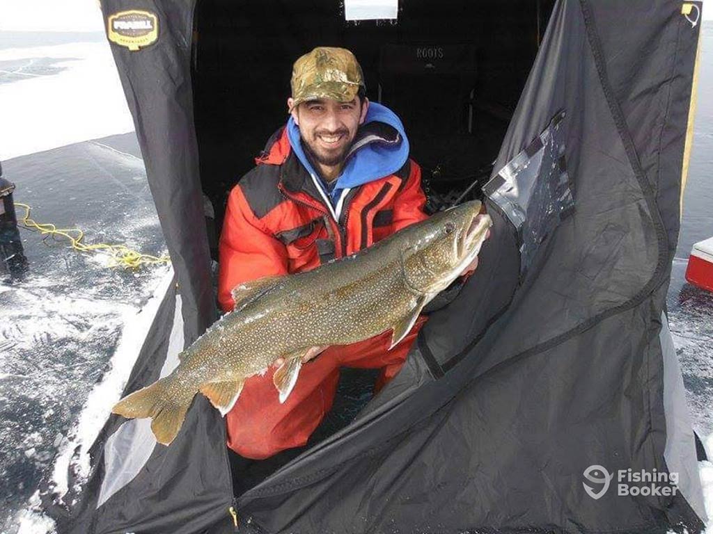 A man crouching in his ice hut, with the zip almost completely undone, holding a Lake Trout caught while ice fishing in Ontario