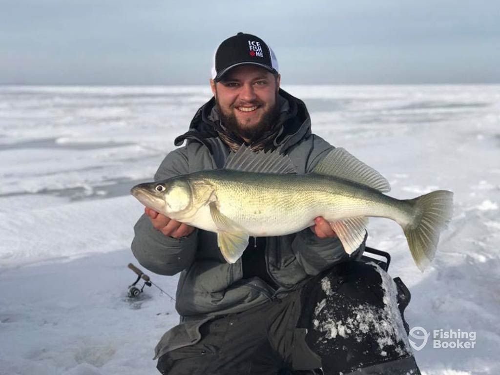 A man standing on a frozen lake in Canada holding a Walleye caught while ice fishing on a clear but cloudy day