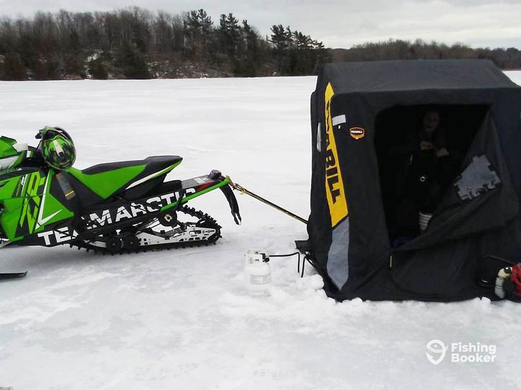 A closeup of a black ice hut used for fishing in Ontario next to a snowmobile on a frozen lake on a cloudy winter's day