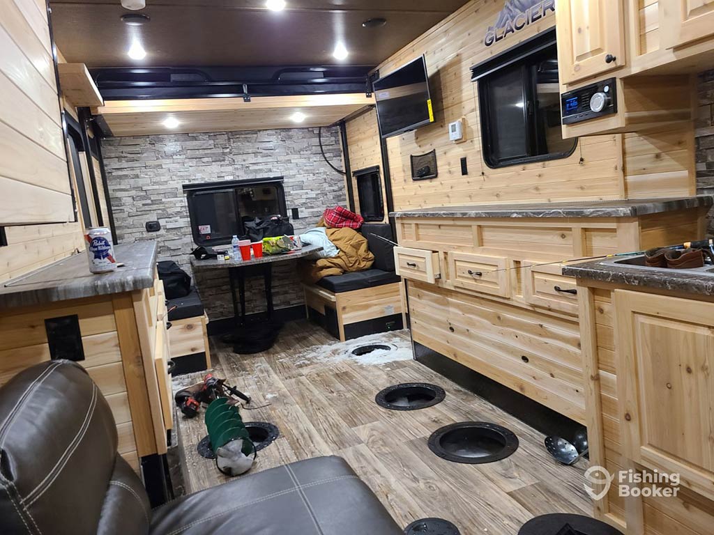 A view of the inside of a fully-equipped ice hut, with holes in the floor ready for ice fishing, along with sofas and a wooden-fitted kitchen area