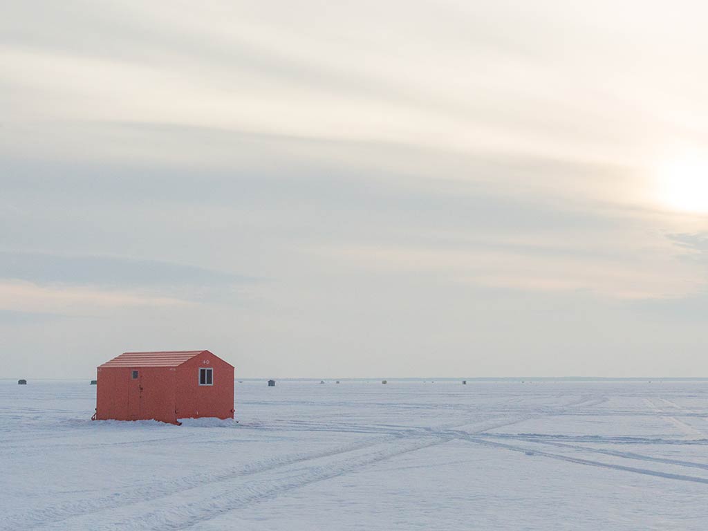 A view across the ice of a frozen Lake Simcoe on a cloudy day during the bright hours towards a red ice hut used for fishing, with some other visible in the far distance