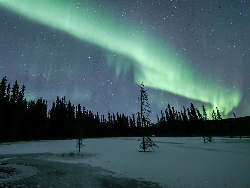 A view of the Northern Lights above a frozen Lake of the Woods in Ontario, Canada, with trees visible in the distance