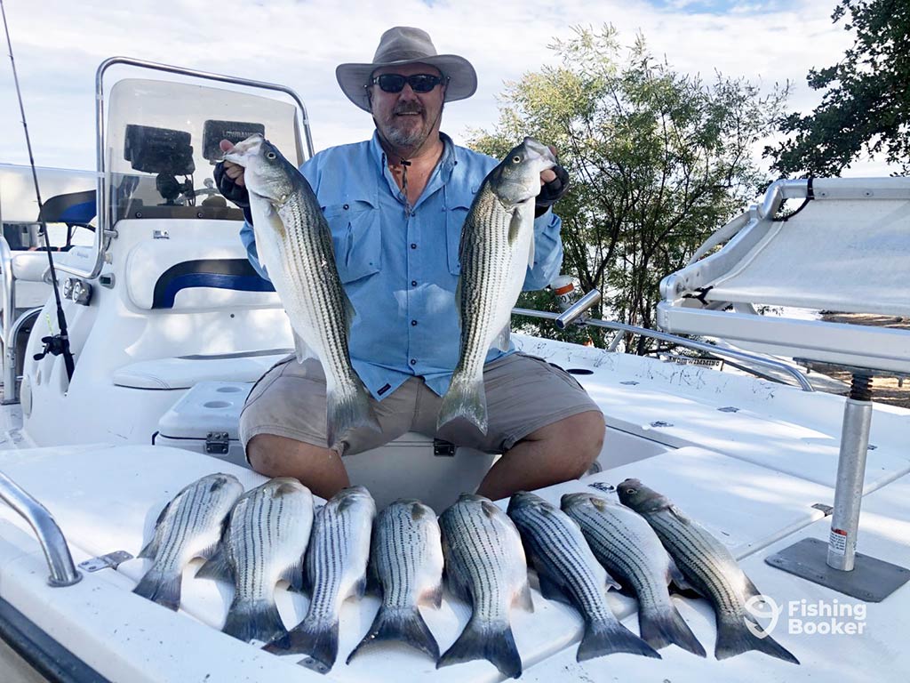 An angler in a hat and sunglasses sitting cross-legged on the bow of a fishing boat after a successful day on the water, holding two Striped Bass and surrounded by eight others laid out around him