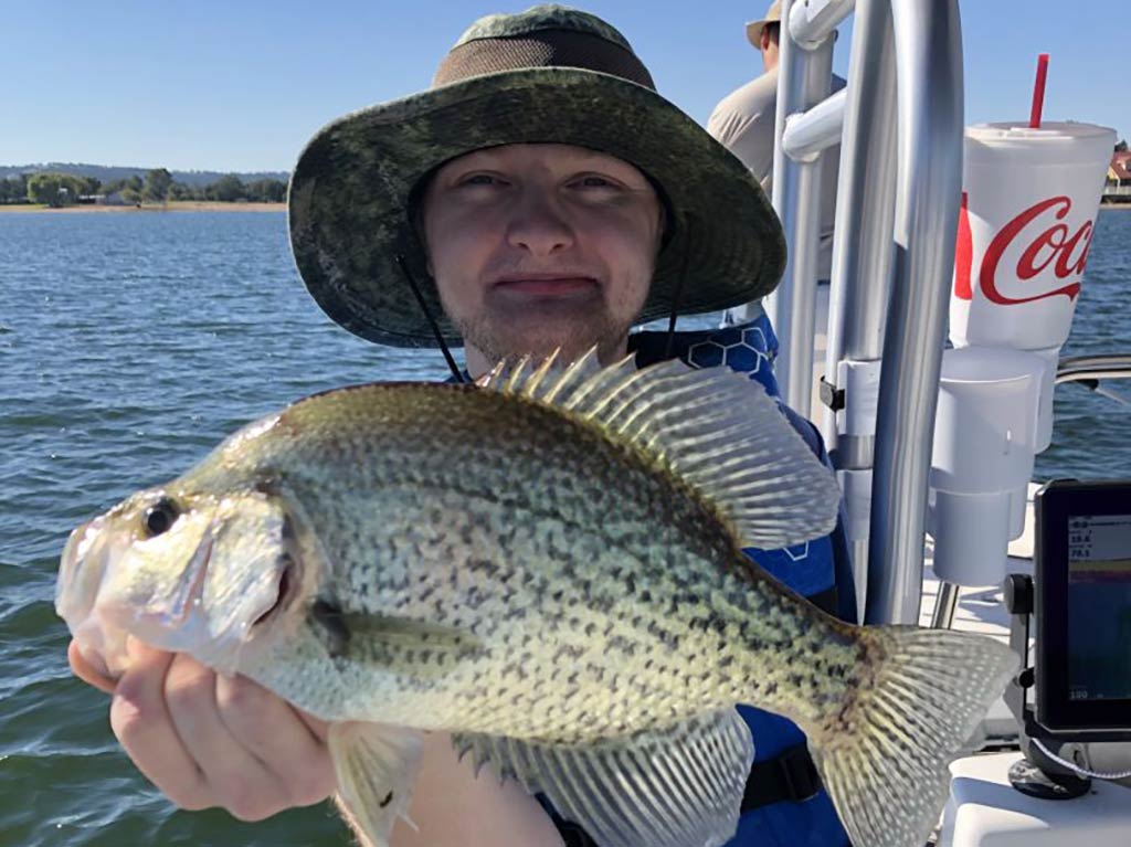 A closeup of a young angler in a hat holding up a Crappie above their face aboard a Lake Livingston fishing charter on a sunny day with water and land visible in the distance