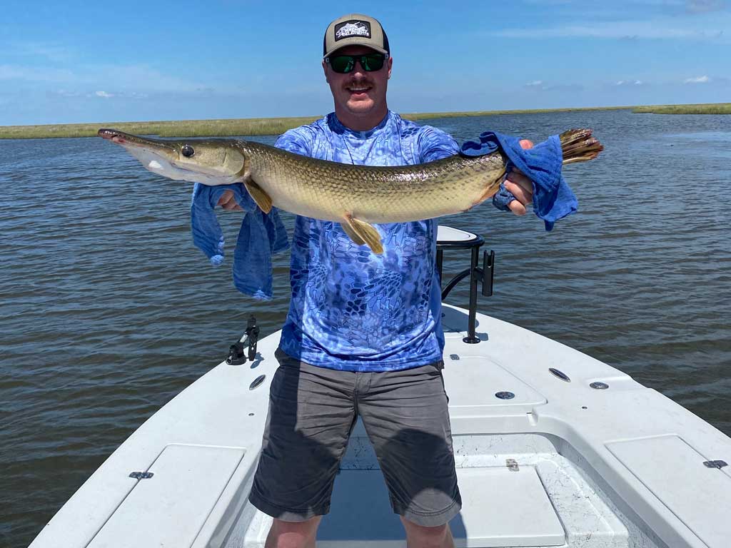 A photo of an angler wearing a cap and a pair of sunglasses while standing on a Louisiana fishing charter boat and posing with Gar