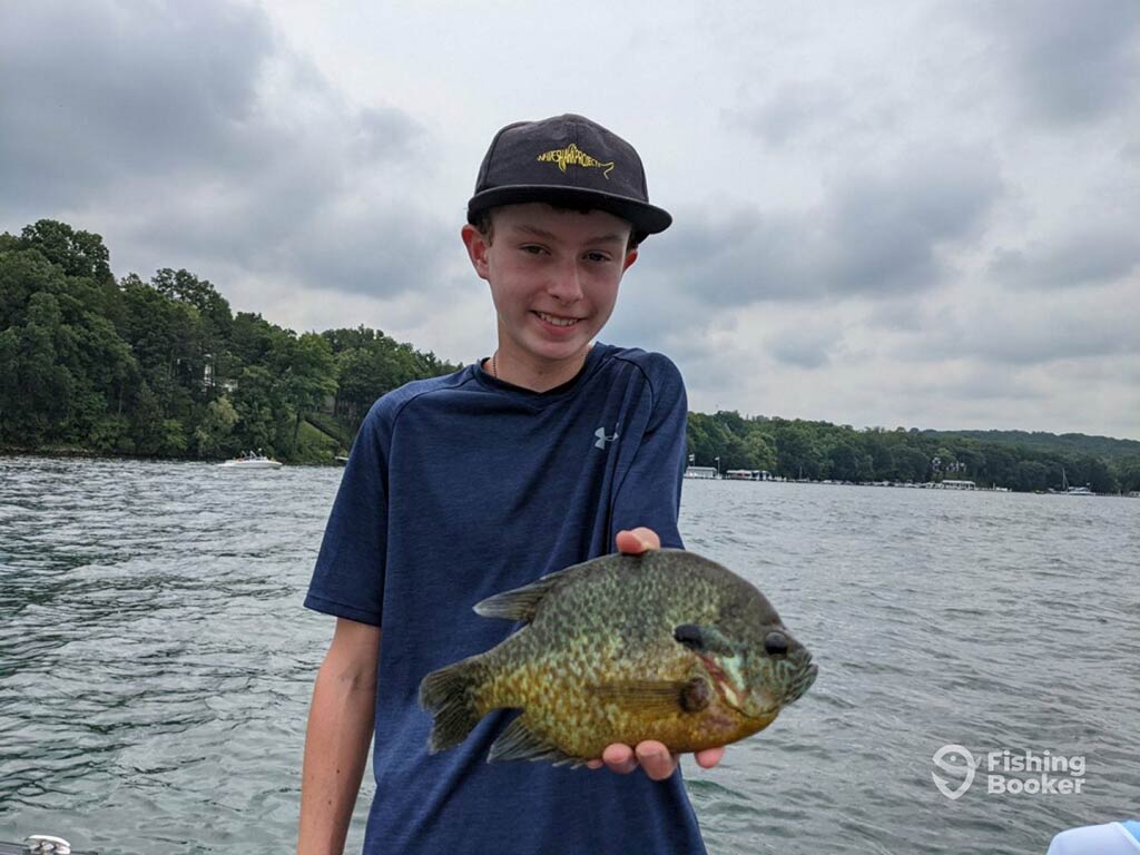 A young boy in a baseball cap standing on a boat and holding a Bluegill fish with the waters of a lake visible behind him on a cloudy day