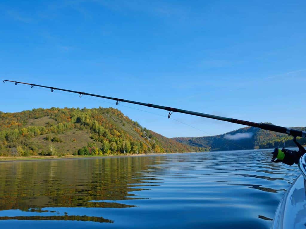 A trolling rod sticks out from the side of a boat on a lake on a sunny day, with land visible ion the distance