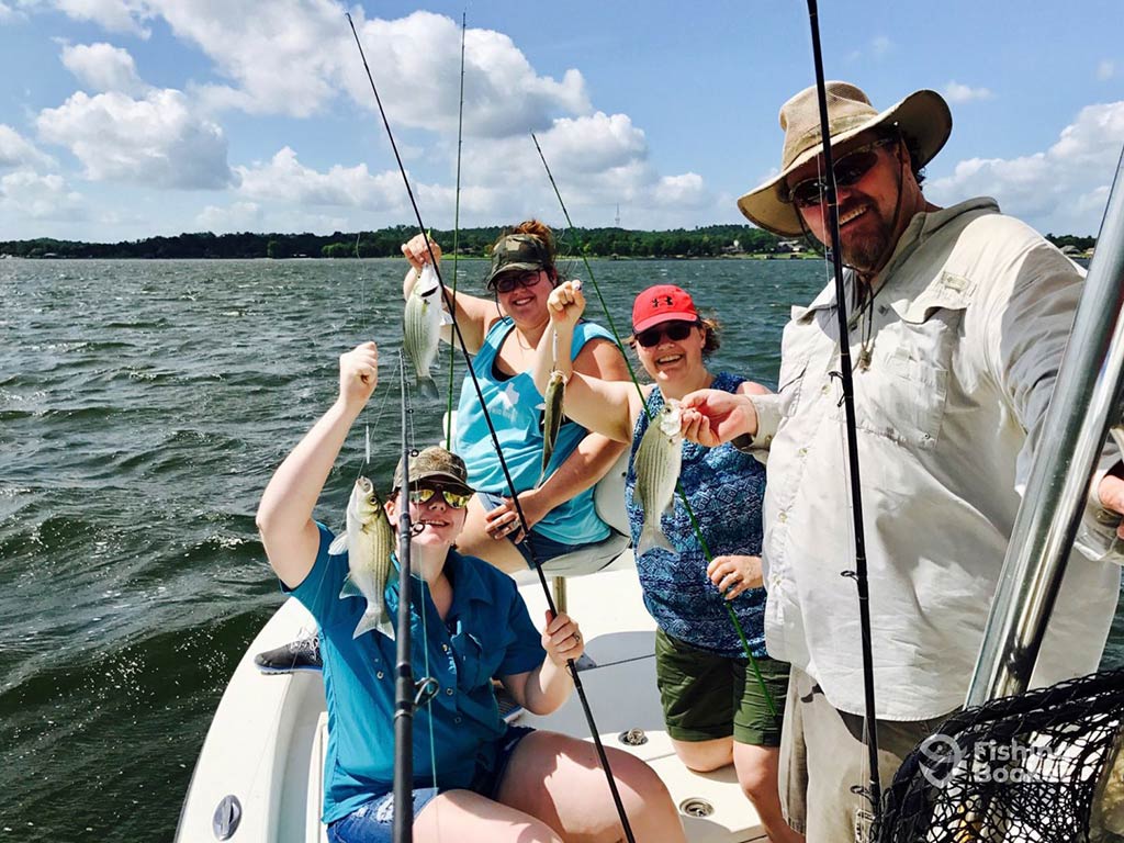 A group of happy anglers aboard a fishing charter in Lake Livingston on a sunny day, with each of them holding a small Striped Bass along with fishing gear, with the water visible behind them and land in the distance