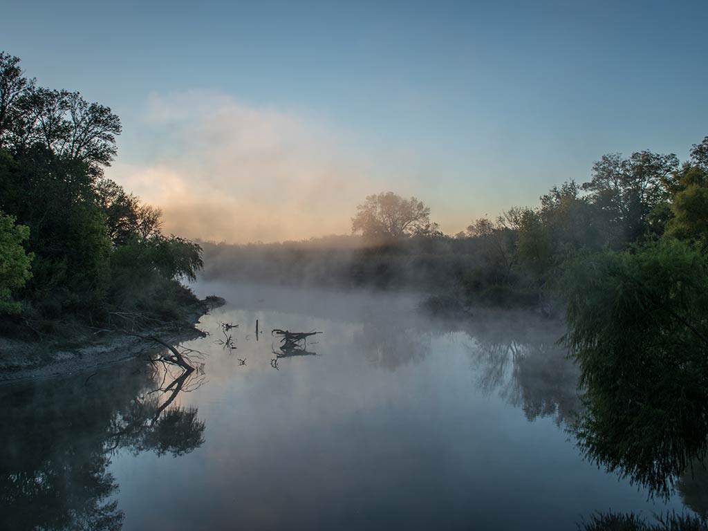 A view along the Trinity River at sunrise, with mist rising up from the lake and the sun visible in the distance behind some clouds